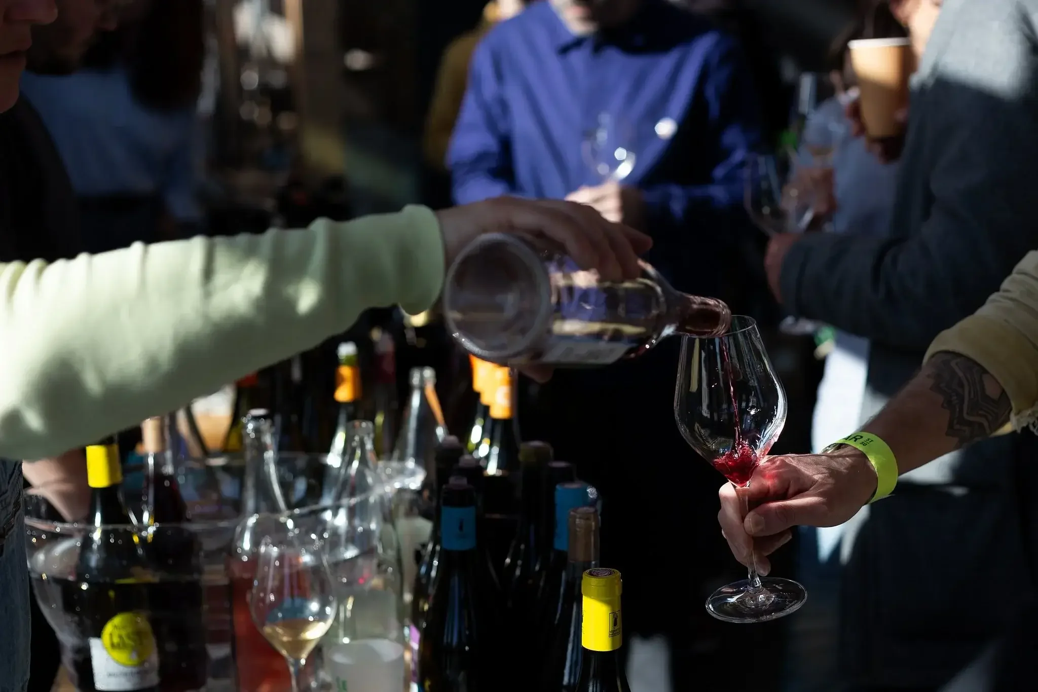 Une personne verse du vin rouge dans un verre à vin à un événement ou à un marché de vin, avec plusieurs bouteilles sur la table.