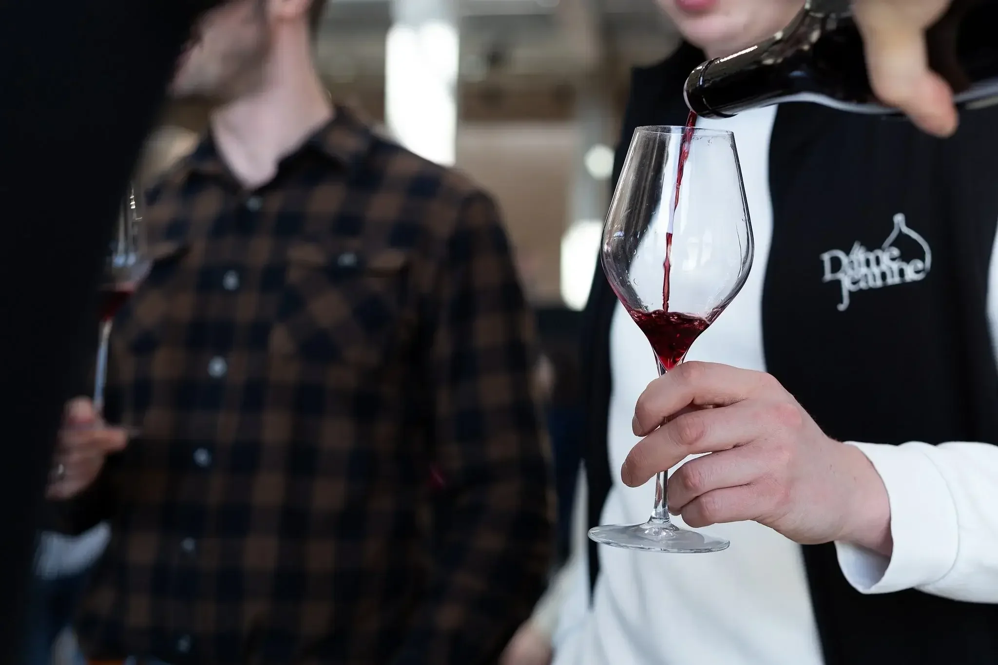 Personne versant du vin rouge dans un verre lors d'une dégustation, avec d'autres personnes en arrière-plan.