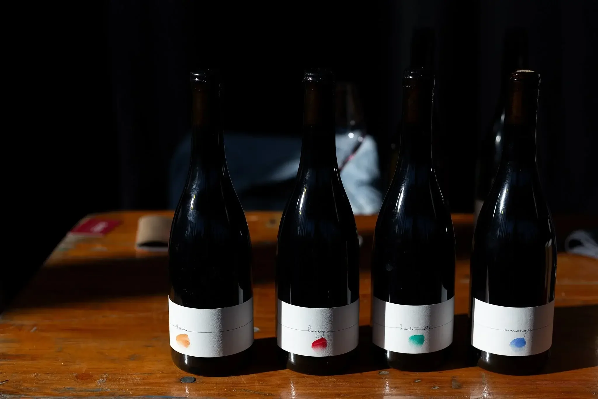 Quatre bouteilles de vin avec étiquettes minimalistes et colorées, alignées sur une table en bois, dans un endroit peu éclairé.