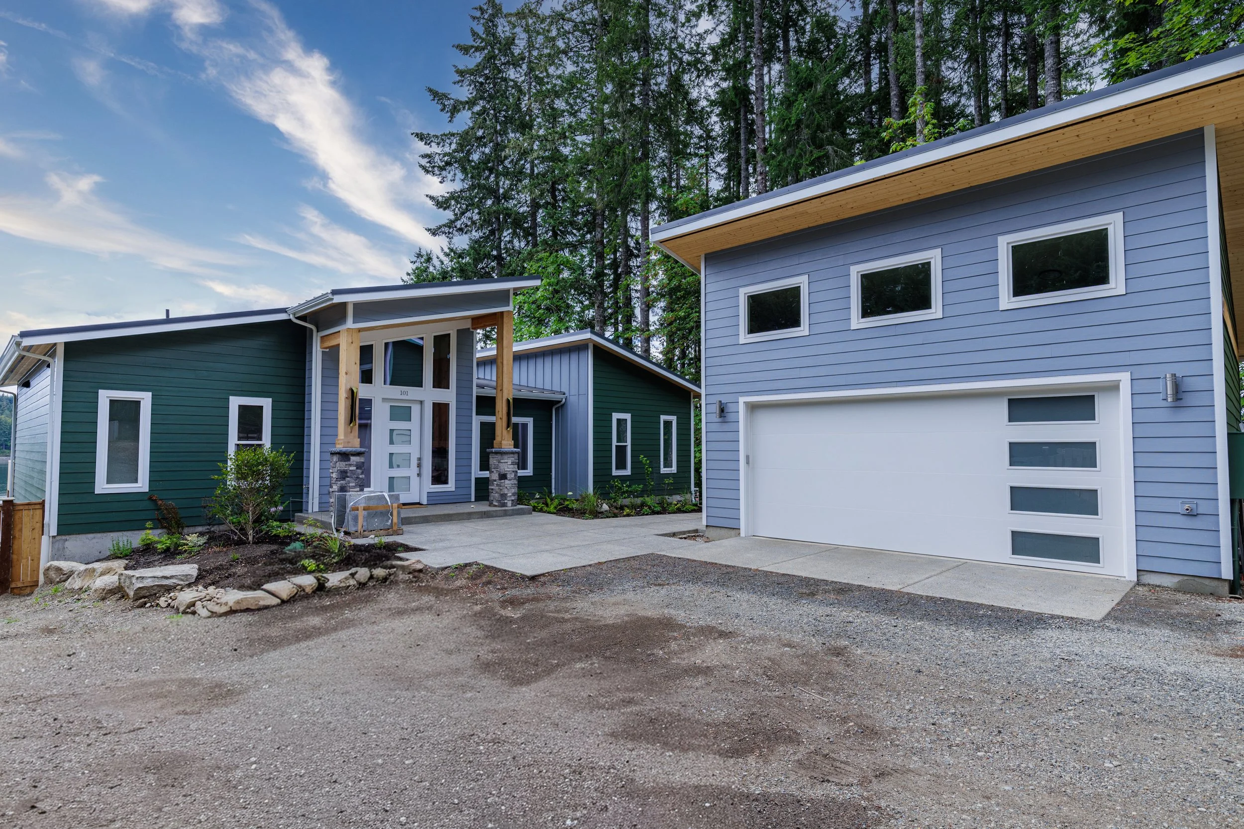 Front exterior of a contemporary custom waterfront home on Pickering Passage in Shelton, WA, featuring deep green and blue-gray siding, offset shed roofs, a wood-framed entry with stone columns, and an attached garage - FoxHead Construction.