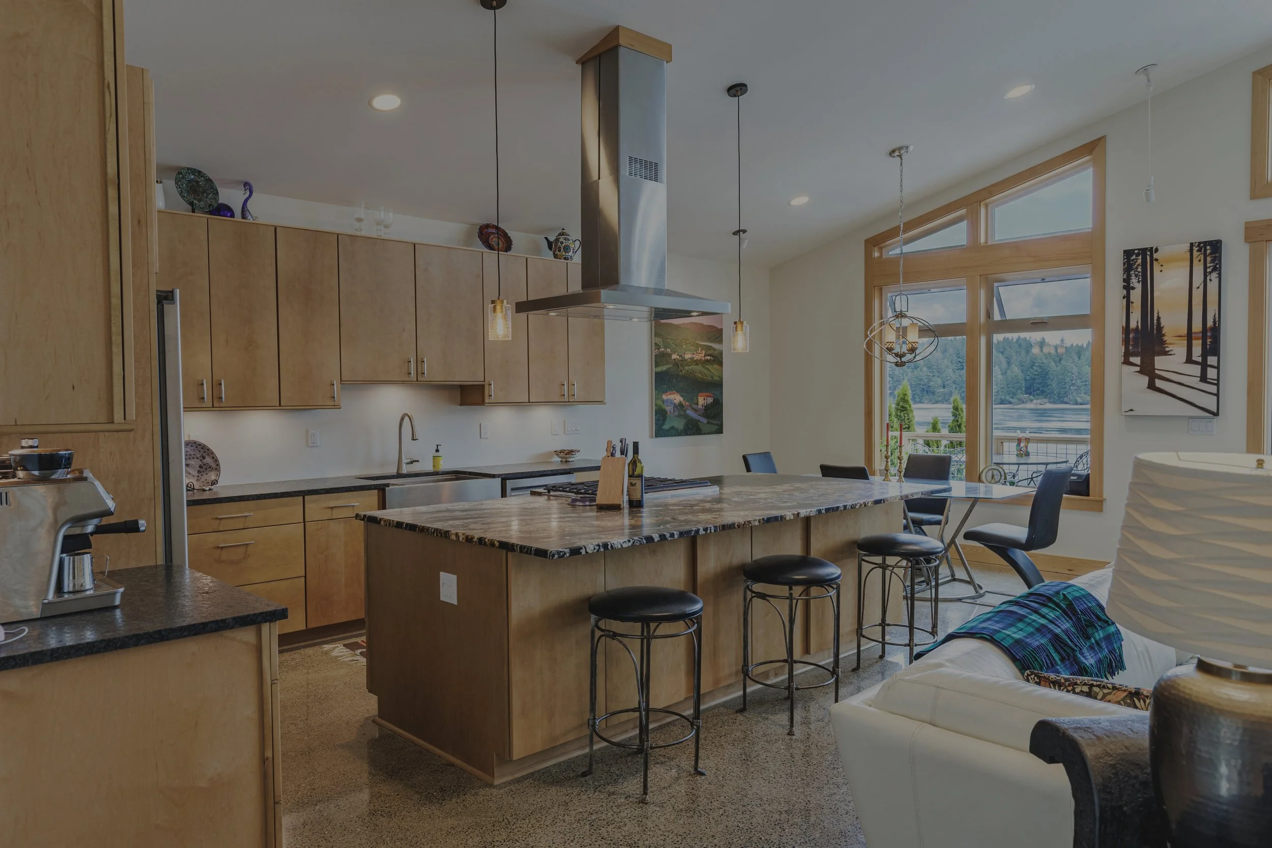 Open kitchen in a Pickering Passage waterfront custom home in Shelton, WA, with natural wood cabinetry, a multicolor granite island, stainless range hood, polished concrete floors, and water views - FoxHead Construction.
