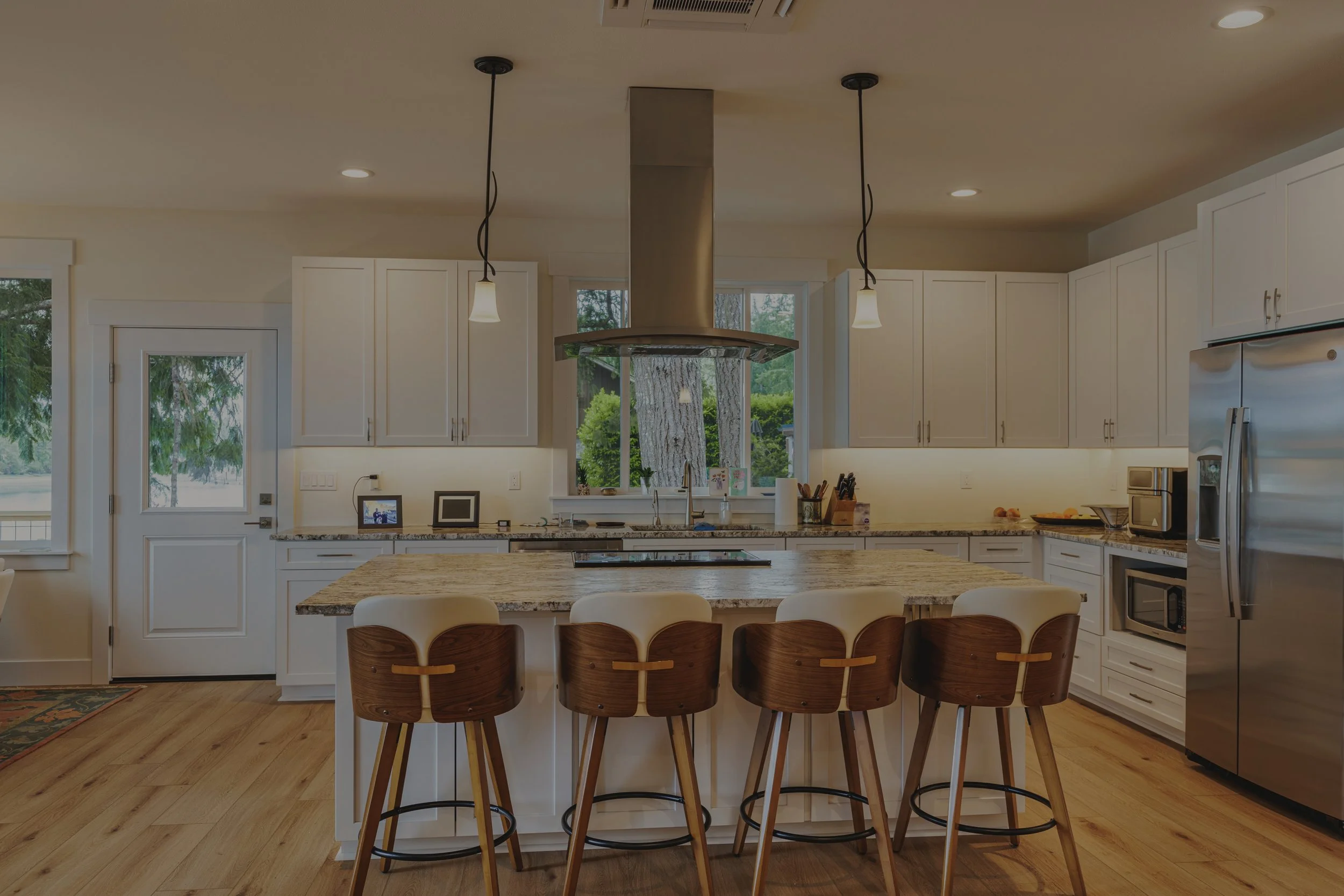 Open kitchen in Margaret’s custom waterfront home on Pickering Passage in Shelton, WA, featuring white cabinetry, a patterned granite island, stainless appliances, wood flooring, and counter seating - FoxHead Construction.