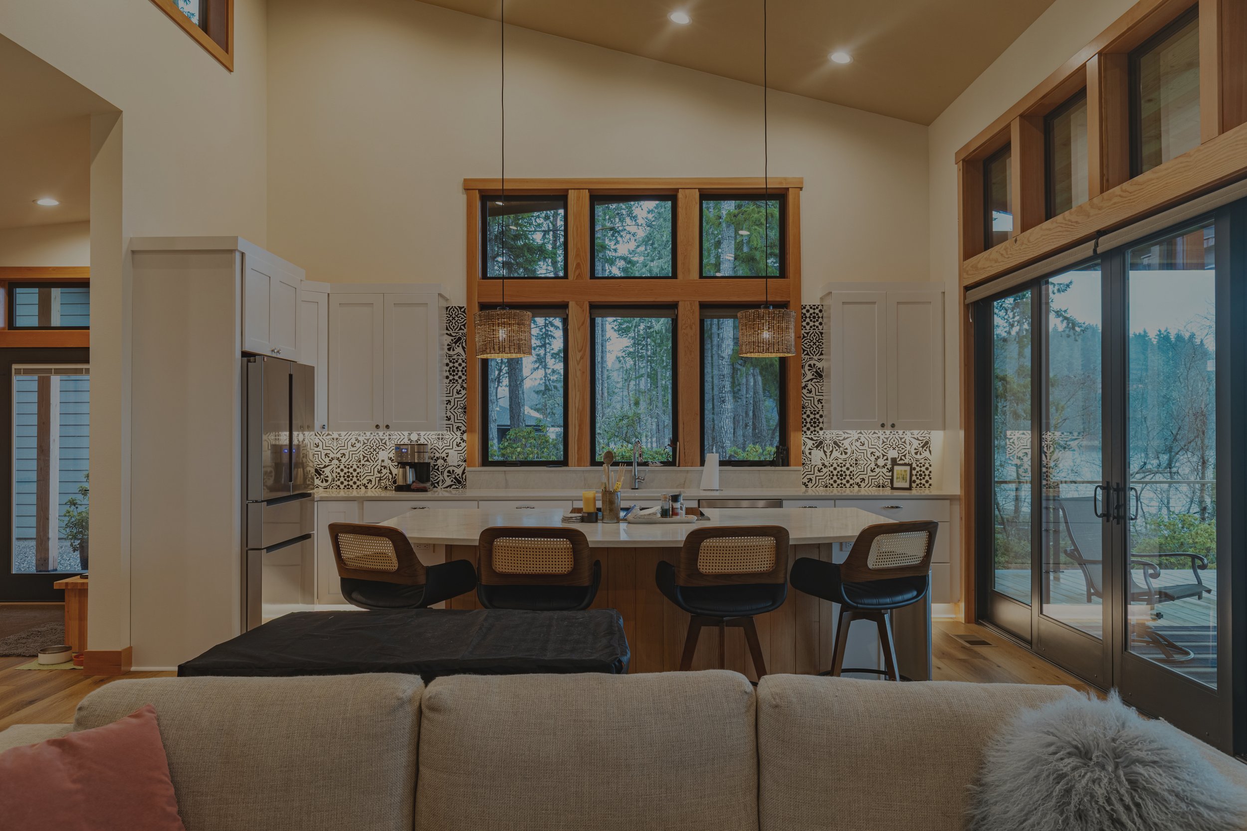 View from the living room into the open-concept kitchen of a modern waterfront custom home on Harstine Island, WA, with large windows showing the evergreen trees and water outside.