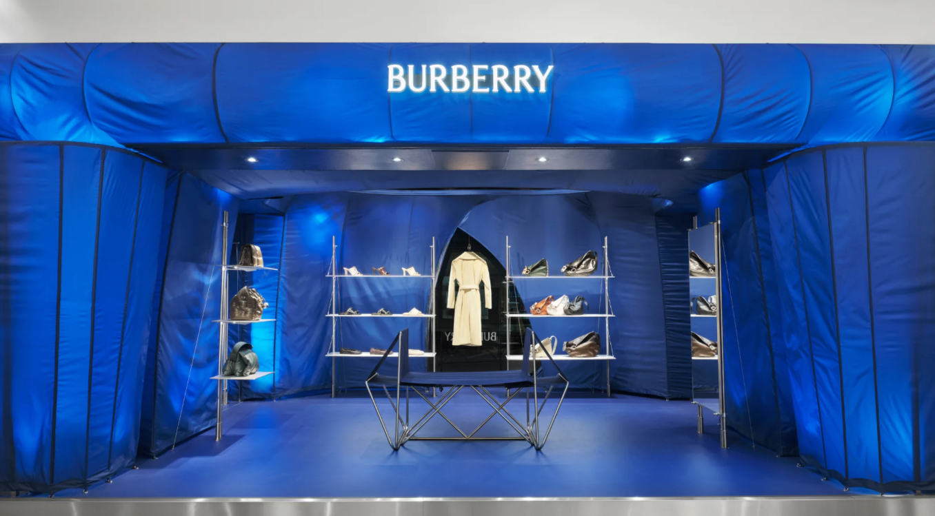 A Burberry pop-up store in Harrods featuring retail display with a blue fabric backdrop, illuminated shelving showcasing handbags, shoes, and accessories, and a white trench coat in the centre.