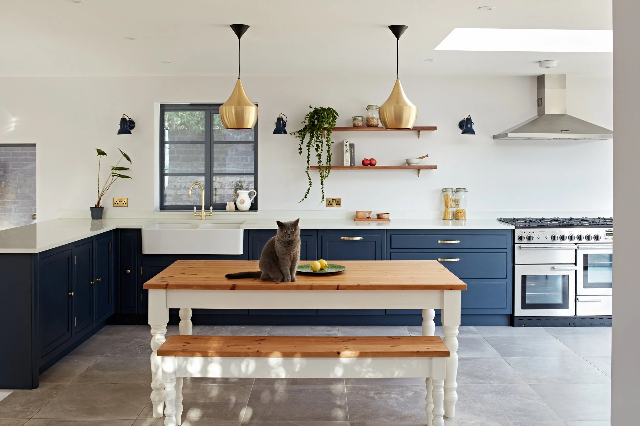 Modern kitchen with navy blue cabinets, white countertops, a farmhouse sink, open shelving with jars and decorative items, a wood dining table with a bench, a gray cat on the table, and a window with natural light.