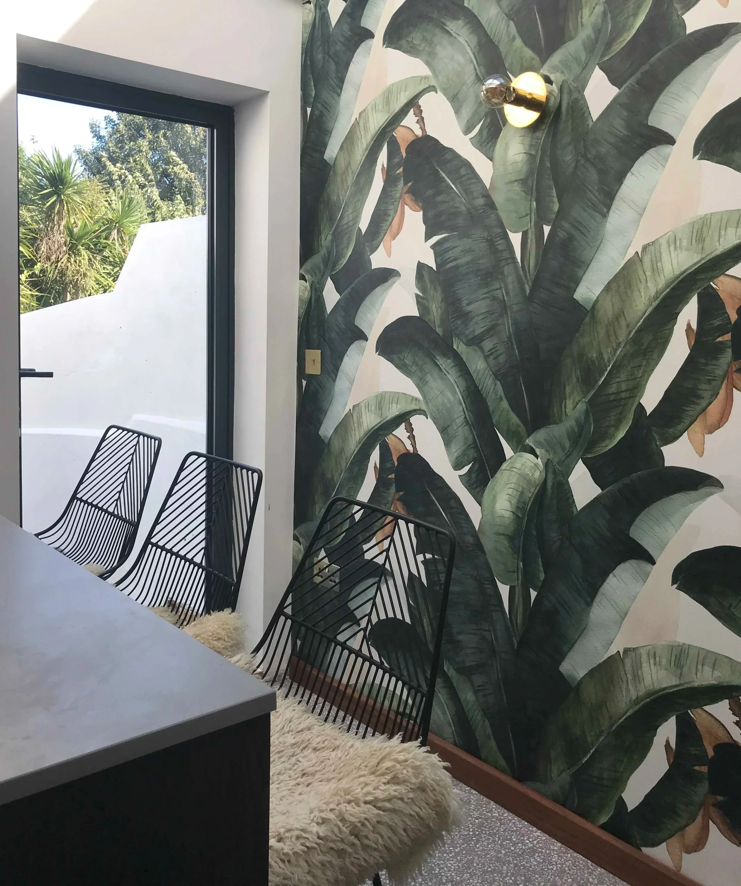 Interior of a modern room featuring a large tropical leaf wallpaper, black wire chairs, a white fluffy cushion, a gray table, and a window with a view of green trees outside.
