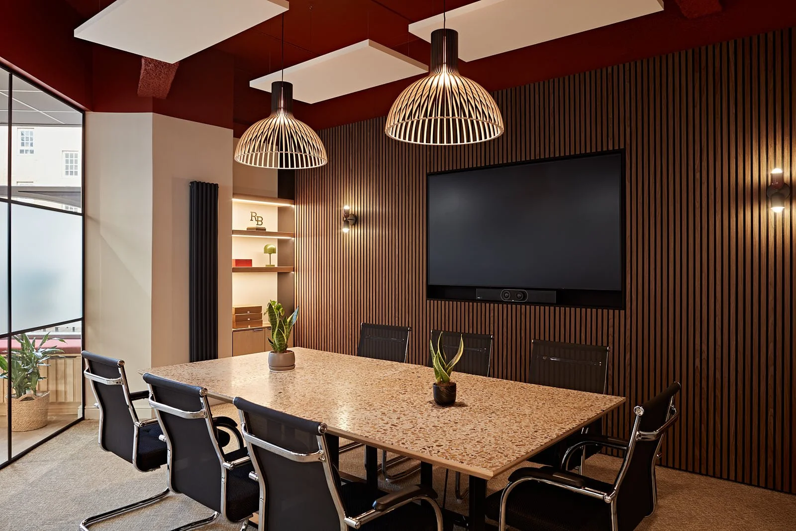Modern conference room with a long table, black chairs, wooden wall paneling, a large screen on the wall, pendant lights, and potted plants