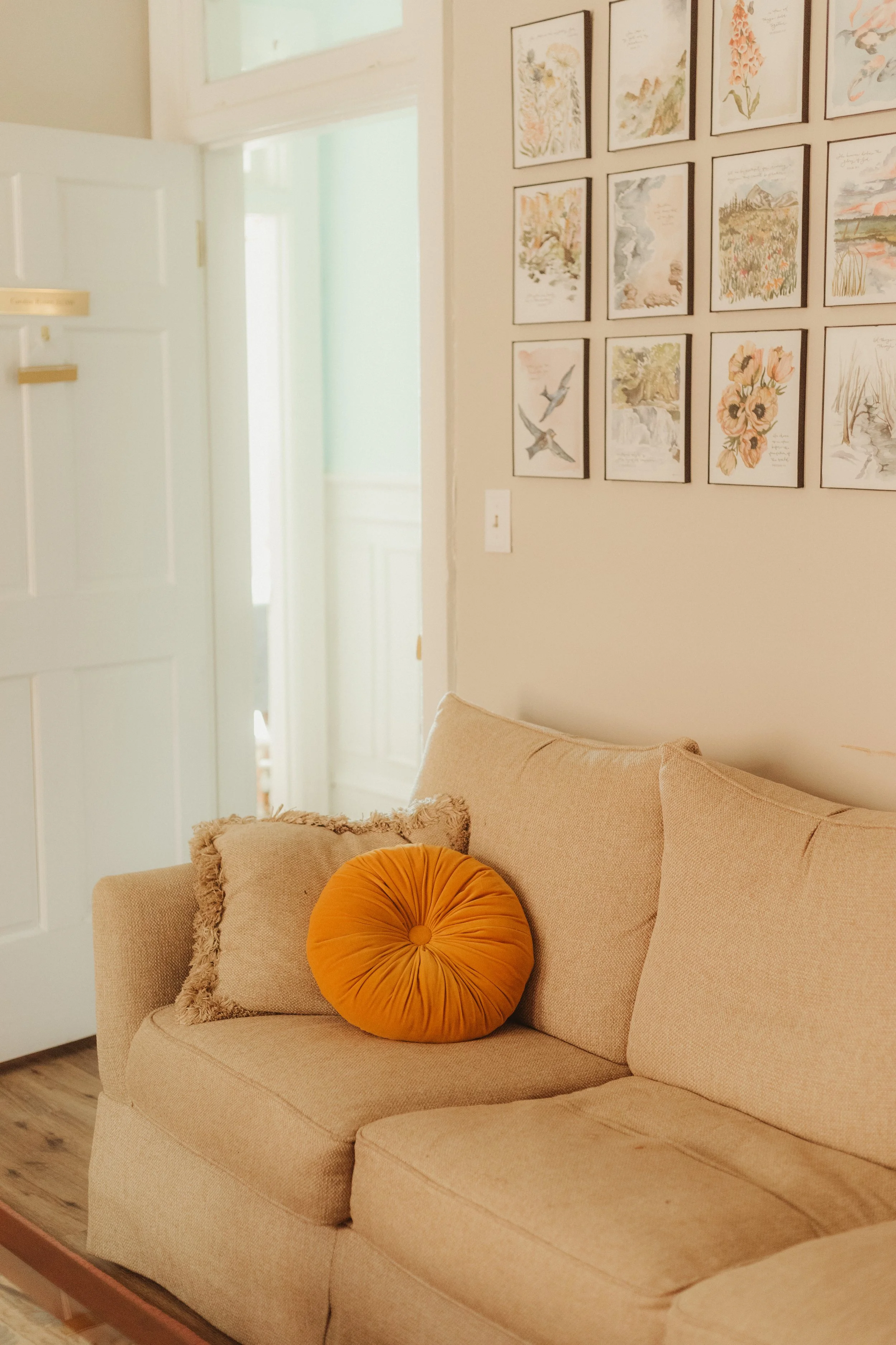 A beige sofa with a round orange cushion and a fuzzy pillow on it. In the background, walls decorated with framed art, and an open door revealing a bright therapy room.