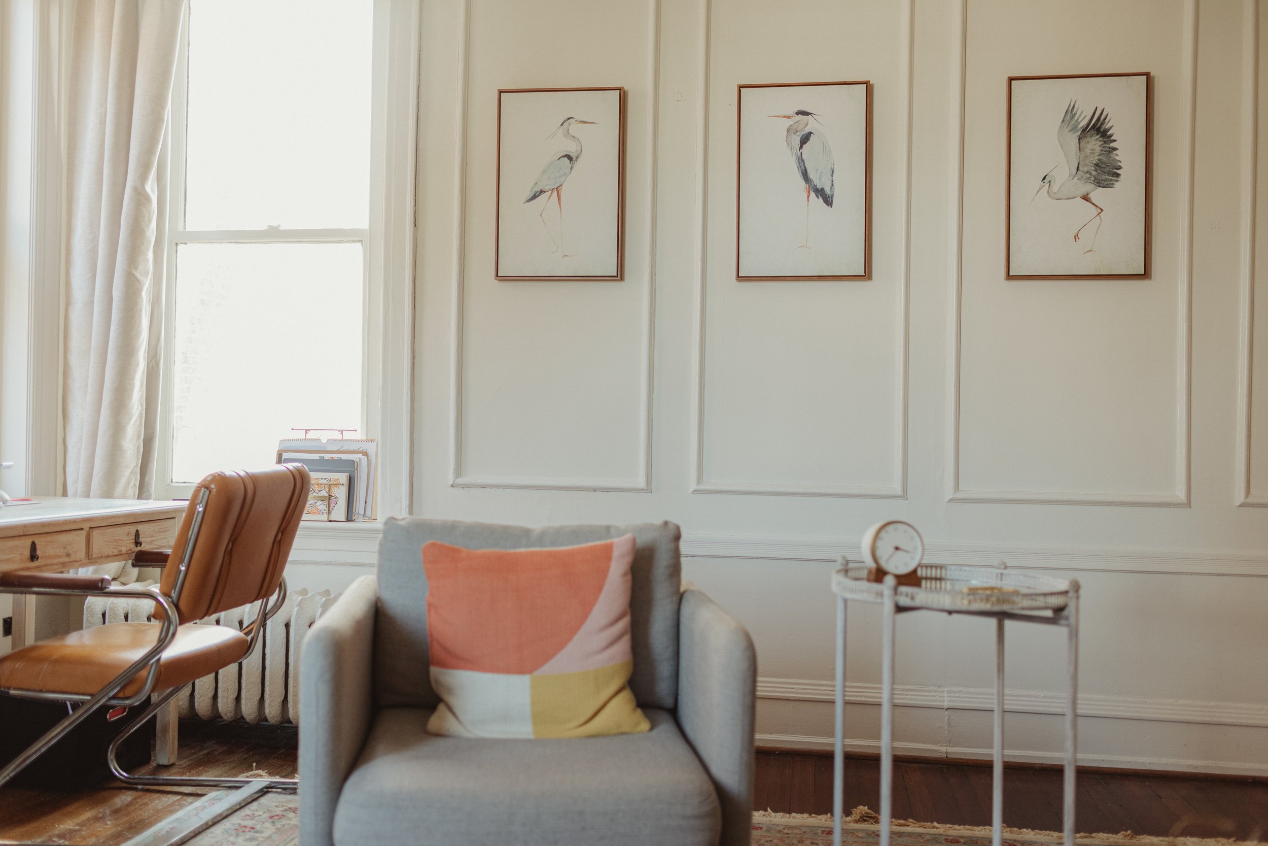 A cozy therapy room with a window, a beige desk chair, a gray armchair with a colorful pillow, a small white side table with a clock, and three framed bird illustrations on the wall.