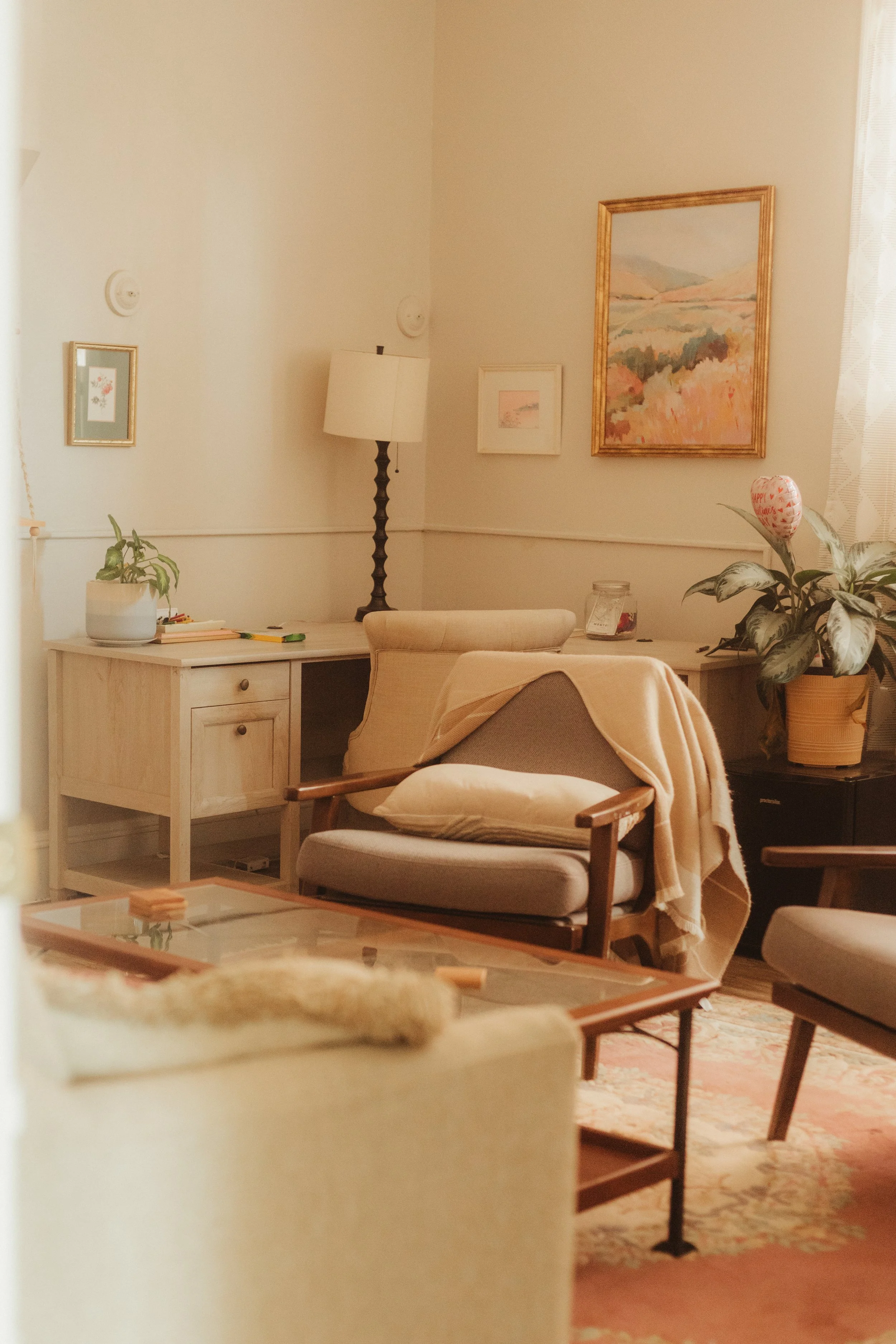 A cozy therapy room corner with a beige armchair, a blanket draped over it, and a pillow. A white desk with a plant and books is in the background.