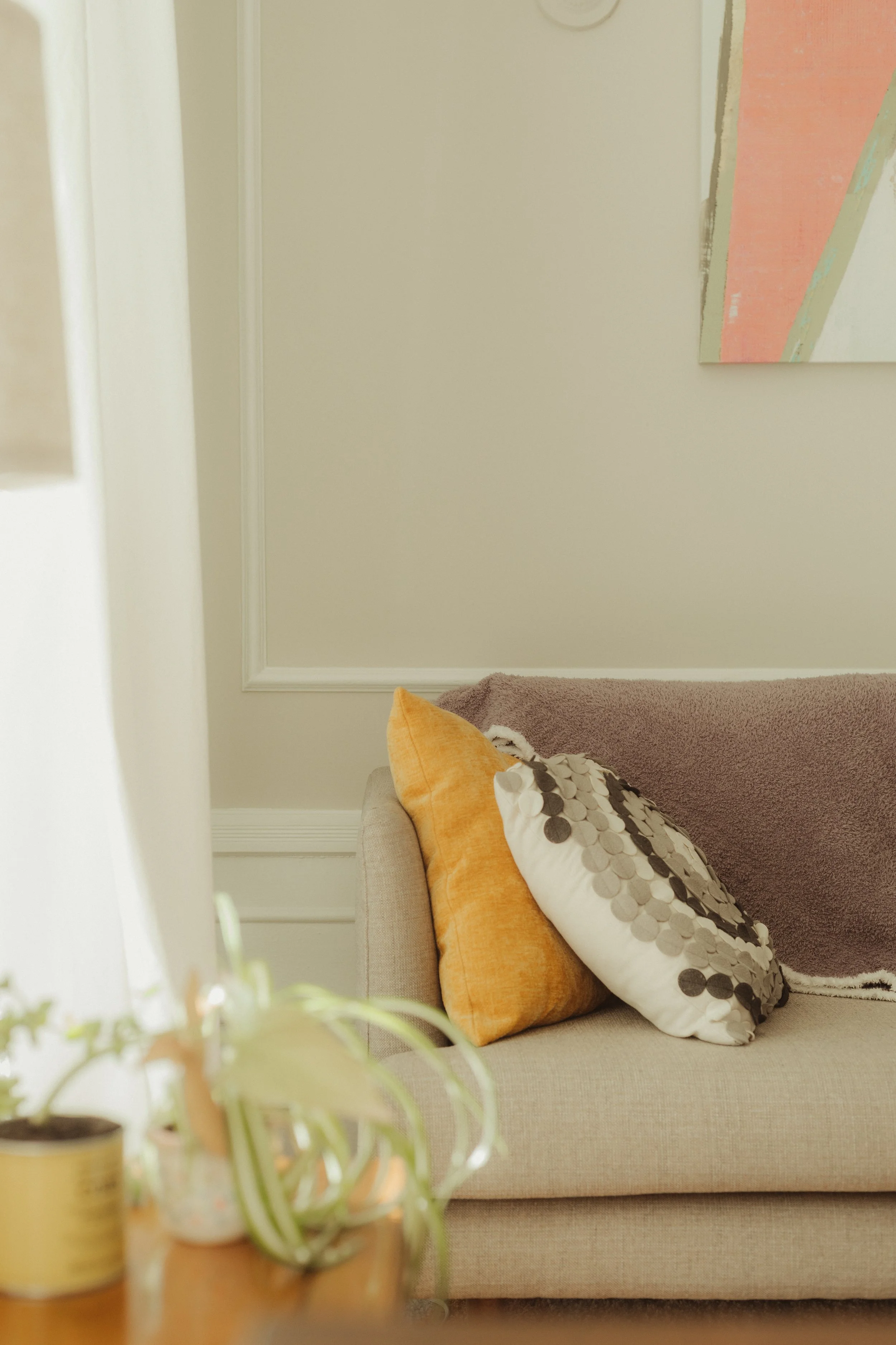 A beige sofa with two throw pillows, one orange and one patterned, in a cozy therapy room with a light-colored wall and abstract art hanging above.