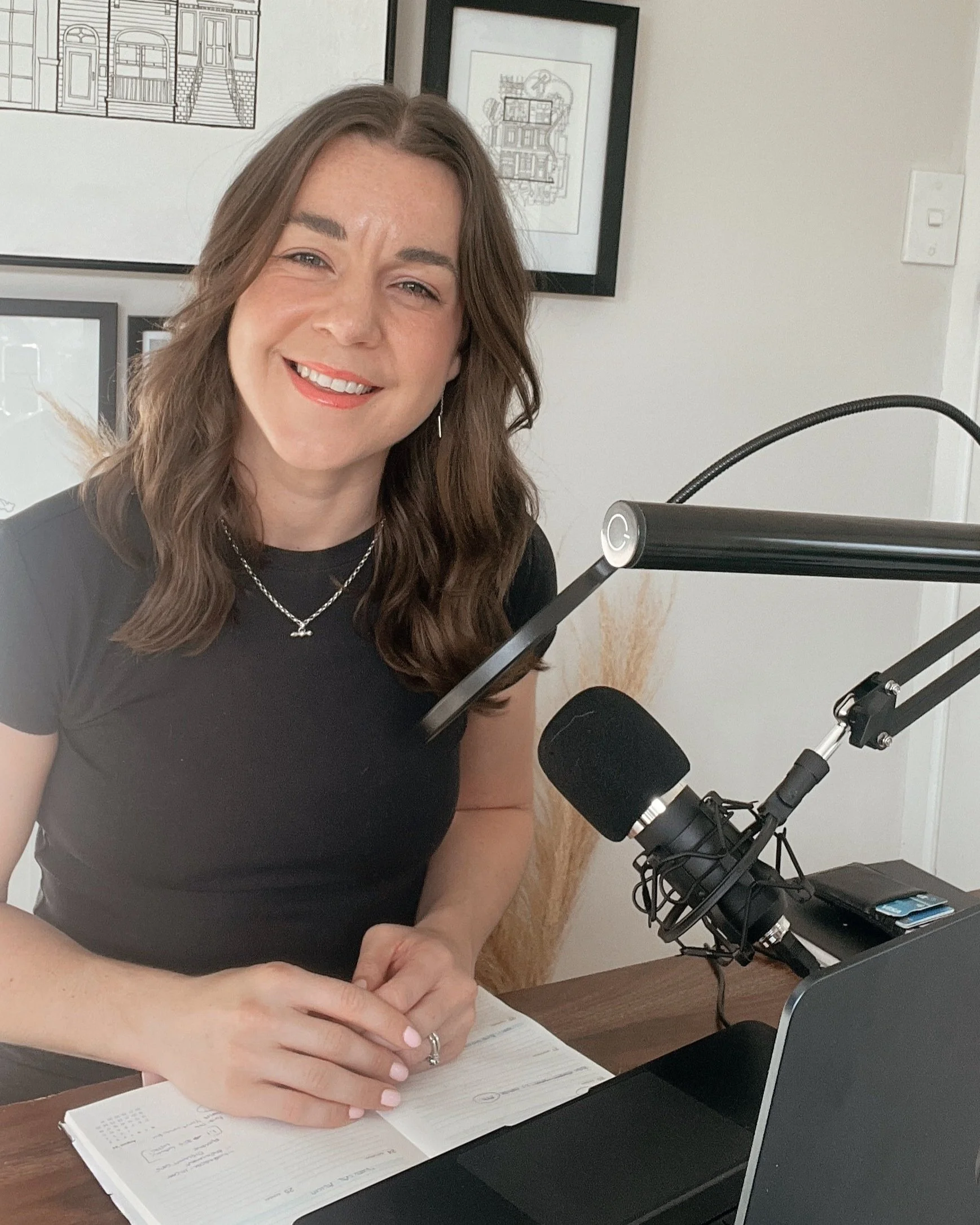 A woman with long brown hair and a black shirt sitting at a desk with a microphone and laptop, smiling into the camera. The background has framed artwork on the wall. She is holding a pen and notebook is open in front of her.