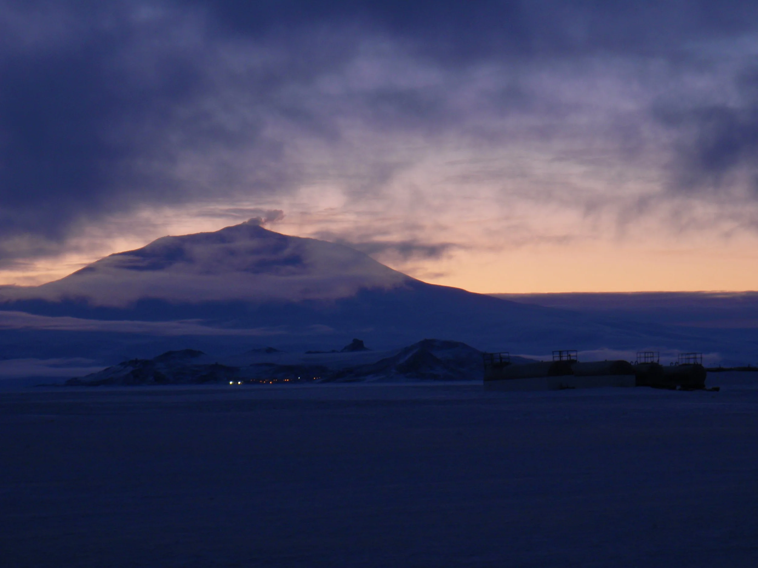 A view of Ross Island from Pegasus Field, during one of the long twilights. Mount Erebus looms in the background over the lights of McMurdo Station. In the foreground are fuel tanks for the airfield. Photo curtesy of Alan R Right, wikimedia