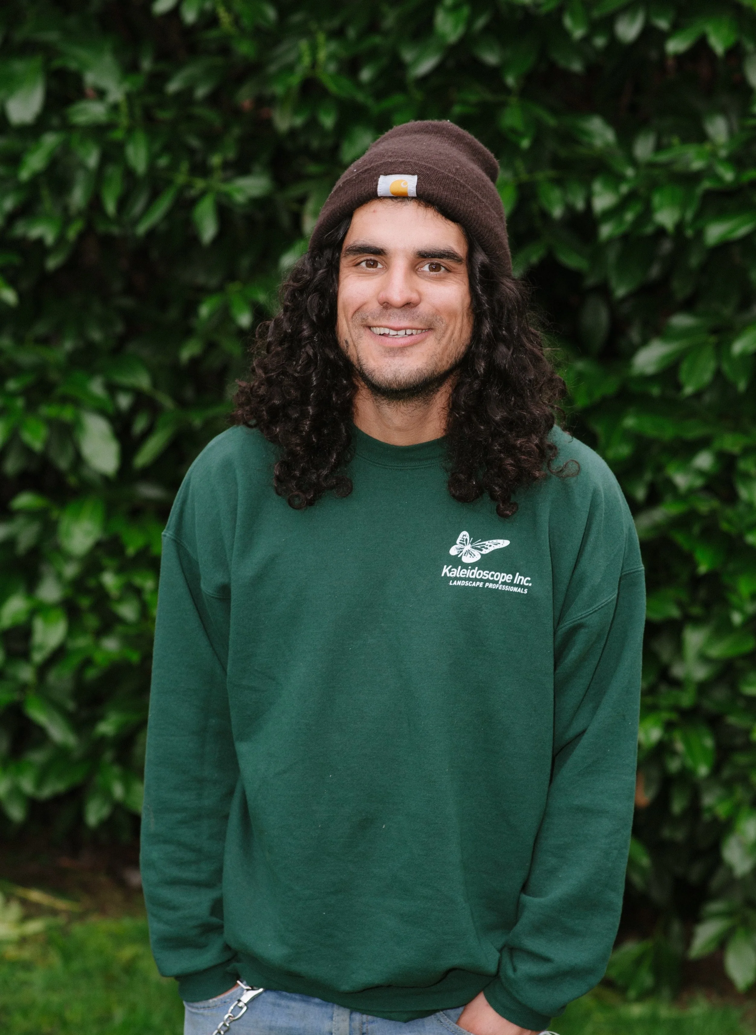 A young man with long, curly dark hair wearing a brown beanie, dark green sweatshirt with 'Kaleidoscope Inc. Landscape Professionals' logo, and light blue jeans, standing outdoors in front of green leafy bushes, smiling at the camera.