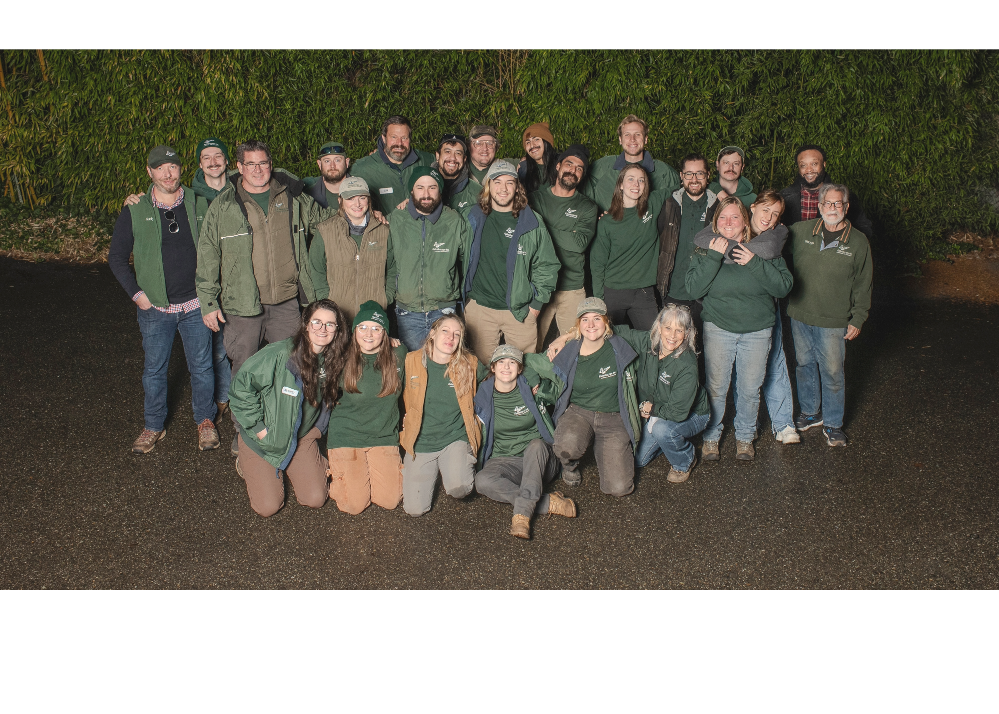 Group of people posing outdoors at night, wearing green and brown jackets, some with branded logos, standing on a dark gravel surface with tall bushes in the background.
