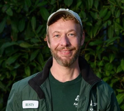 A man with a beard and mustache, smiling, wearing a gray cap, a green jacket with a name tag that says 'Rusty', and a green shirt.