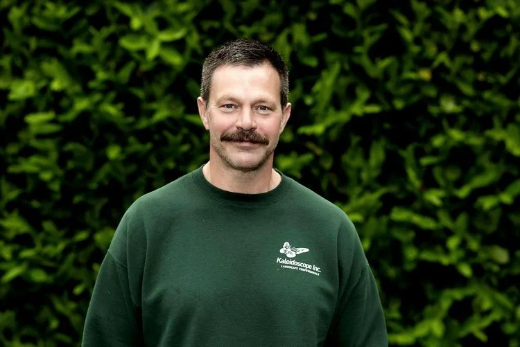 Man with dark hair and mustache wearing a green sweatshirt with a logo, standing outdoors in front of leafy green background.