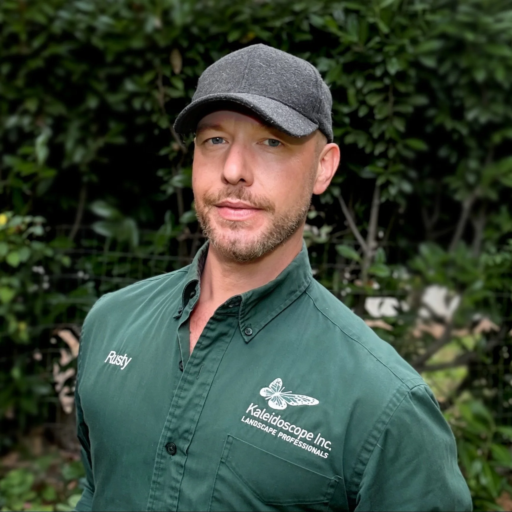 A man with a beard and mustache, smiling, wearing a gray cap, a green jacket with a name tag that says 'Rusty', and a green shirt.
