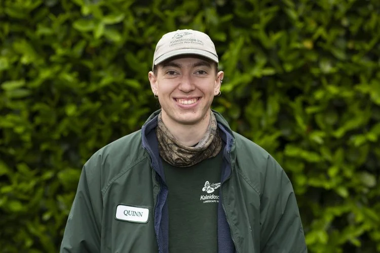 A young man wearing a beige cap, green jacket, and a dark green shirt stands outdoors with a leafy green background, smiling.
