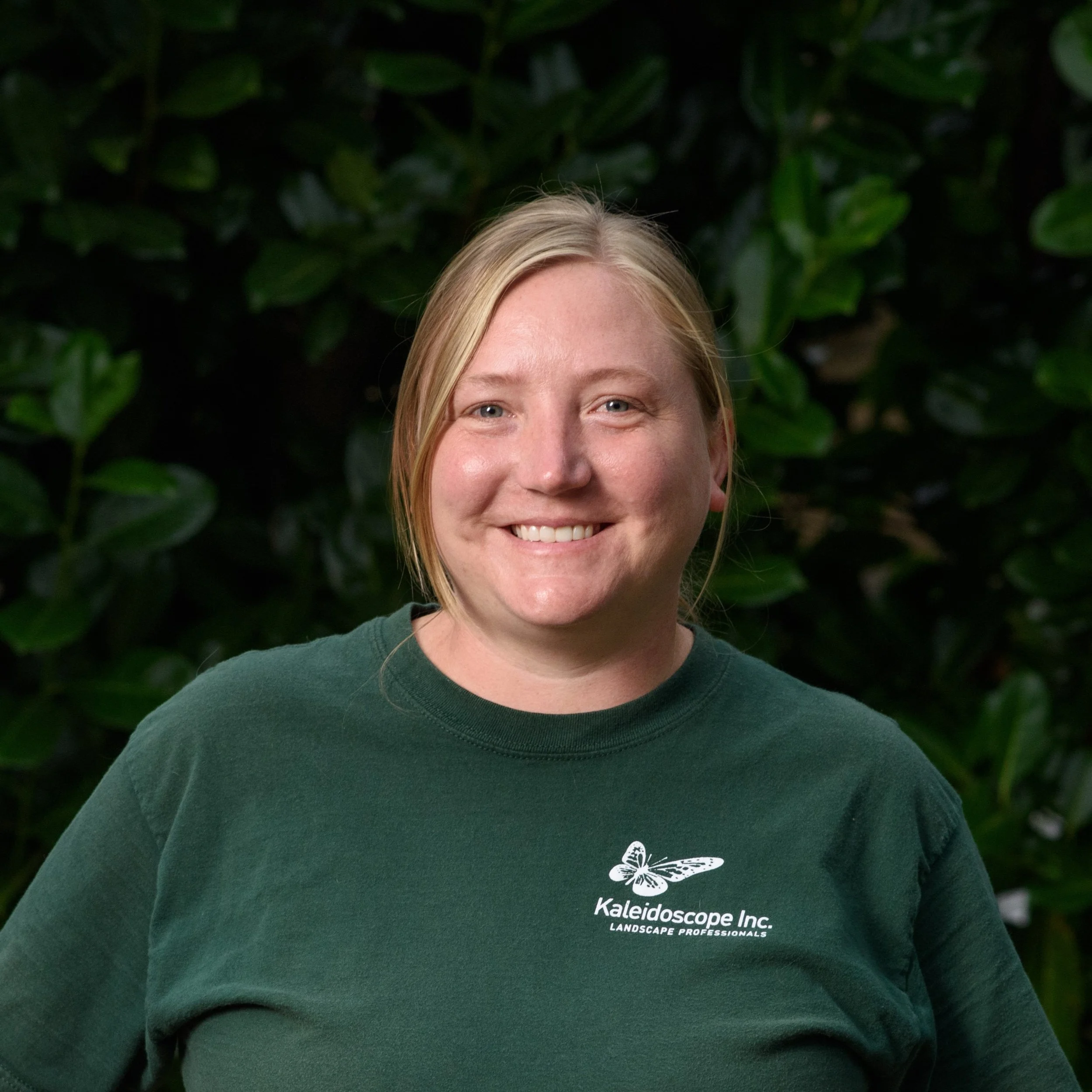 A woman with blonde hair smiling, wearing a dark green shirt with a logo that says Kaleidoscope Inc. Landscape Professionals, standing in front of green foliage.