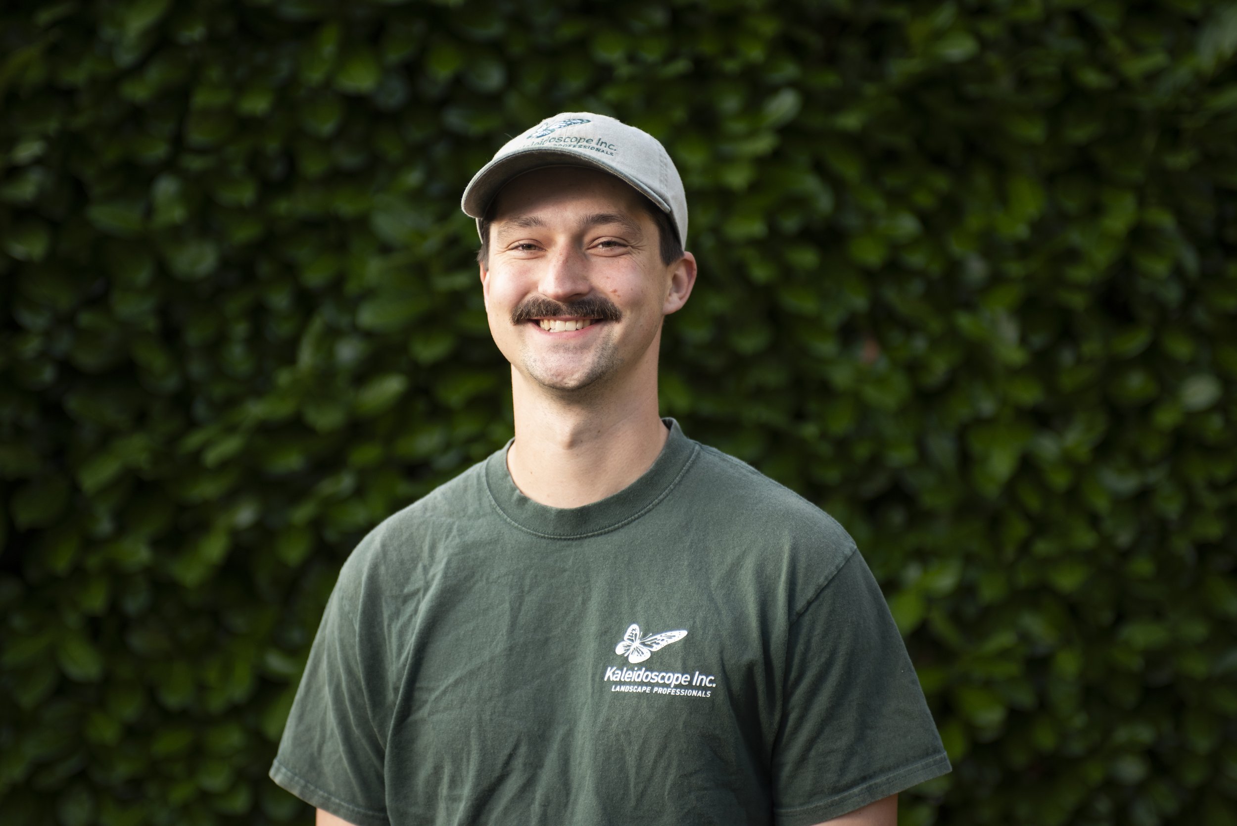 A young man smiling outdoors, wearing a light-colored cap and a dark green T-shirt with a company logo.