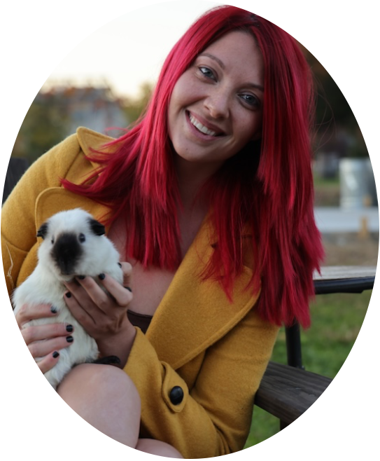 A professional headshot of Breanna R., a pet sitter in Denver, smiling and holding a small white and black guinea pig. She has bright red hair and is wearing a mustard yellow coat.