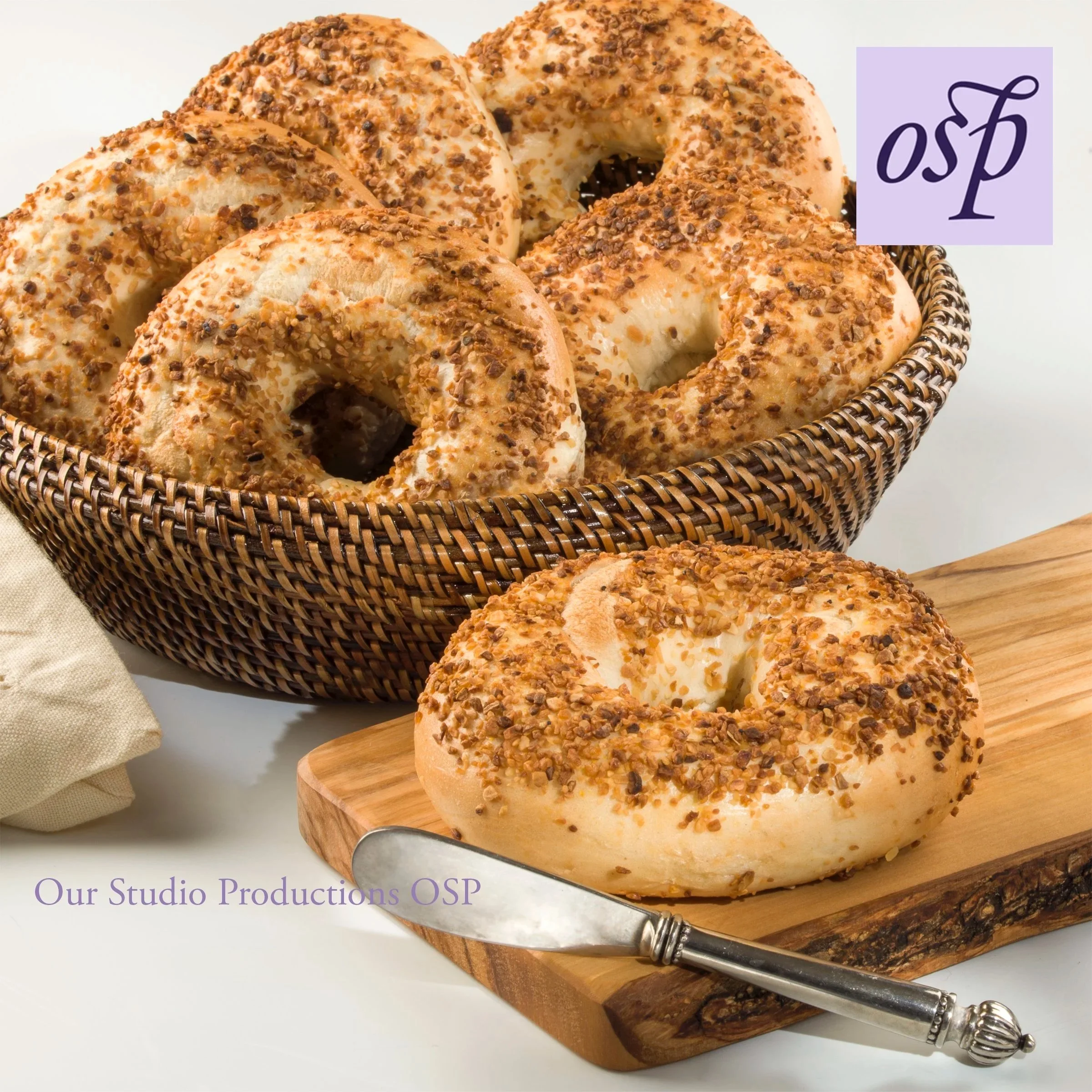 Bagels in a basket, with a butter knife placed alongside of a wood cutting board.