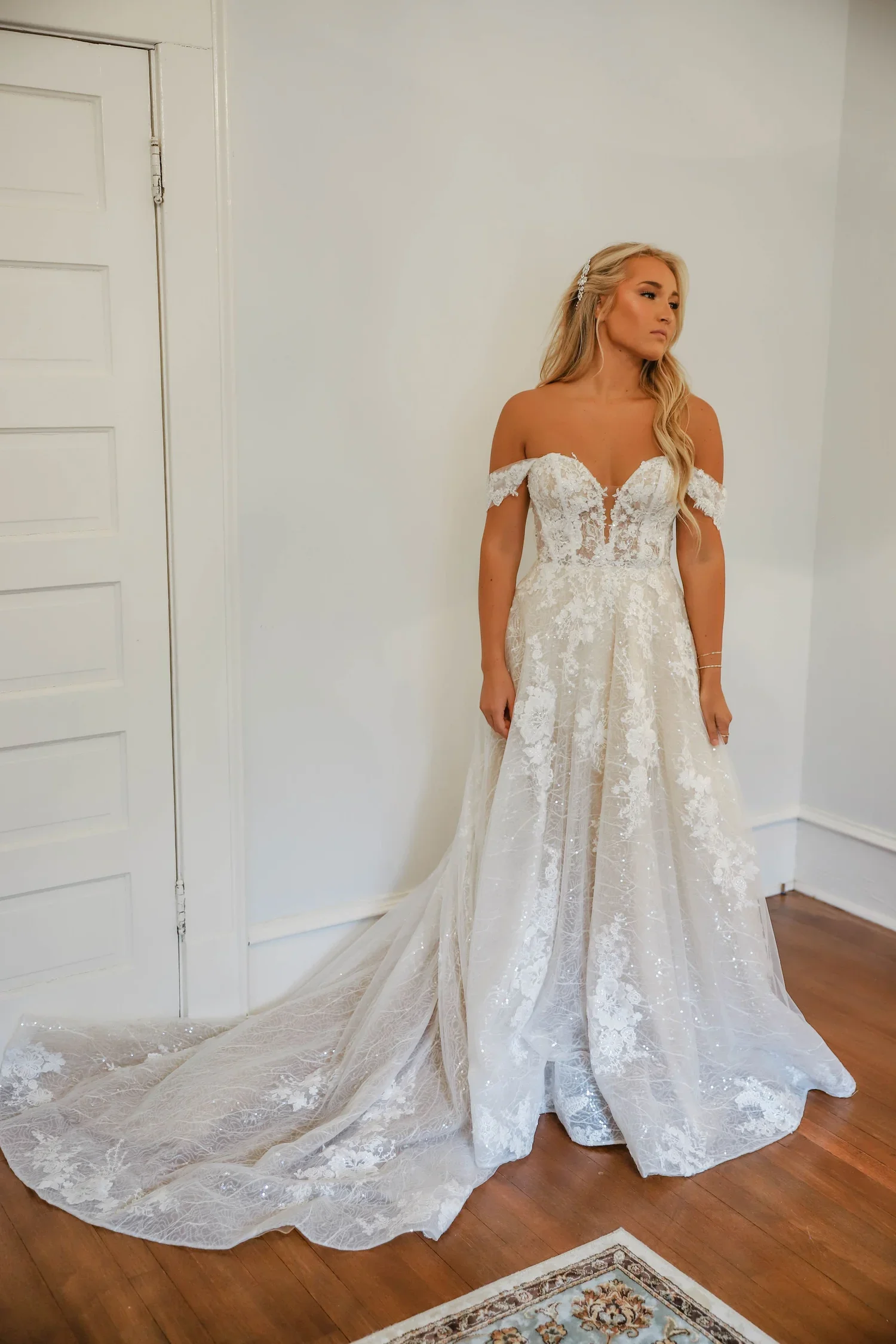 Bride wearing an off-shoulder lace A-line wedding gown with a flowing train and floral applique details, standing in a softly lit room at Atlanta Street Bridal Company in McDonough, GA