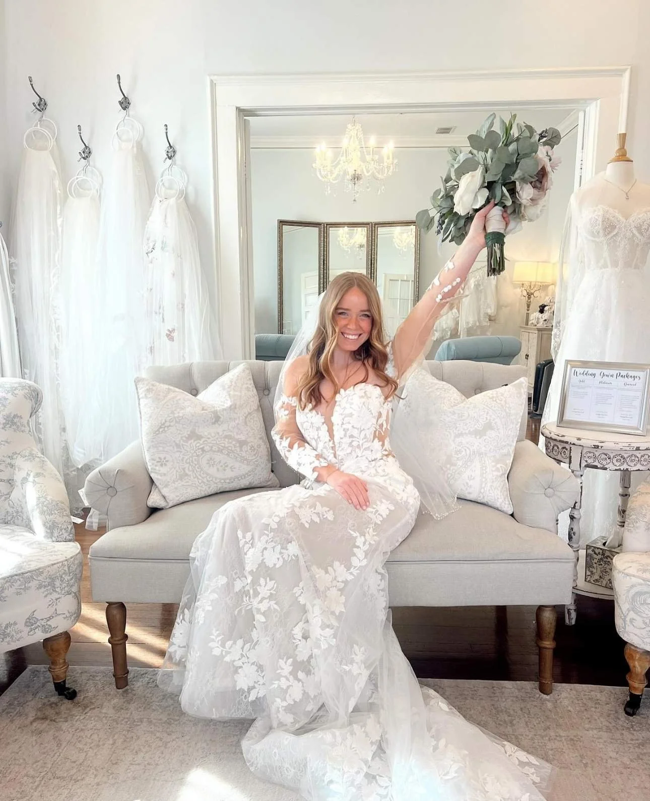 Bride celebrating in a lace wedding dress with long sleeves, seated on a sofa and holding a bouquet inside the Atlanta Street Bridal showroom, surrounded by veils, mirrors, and elegant décor.