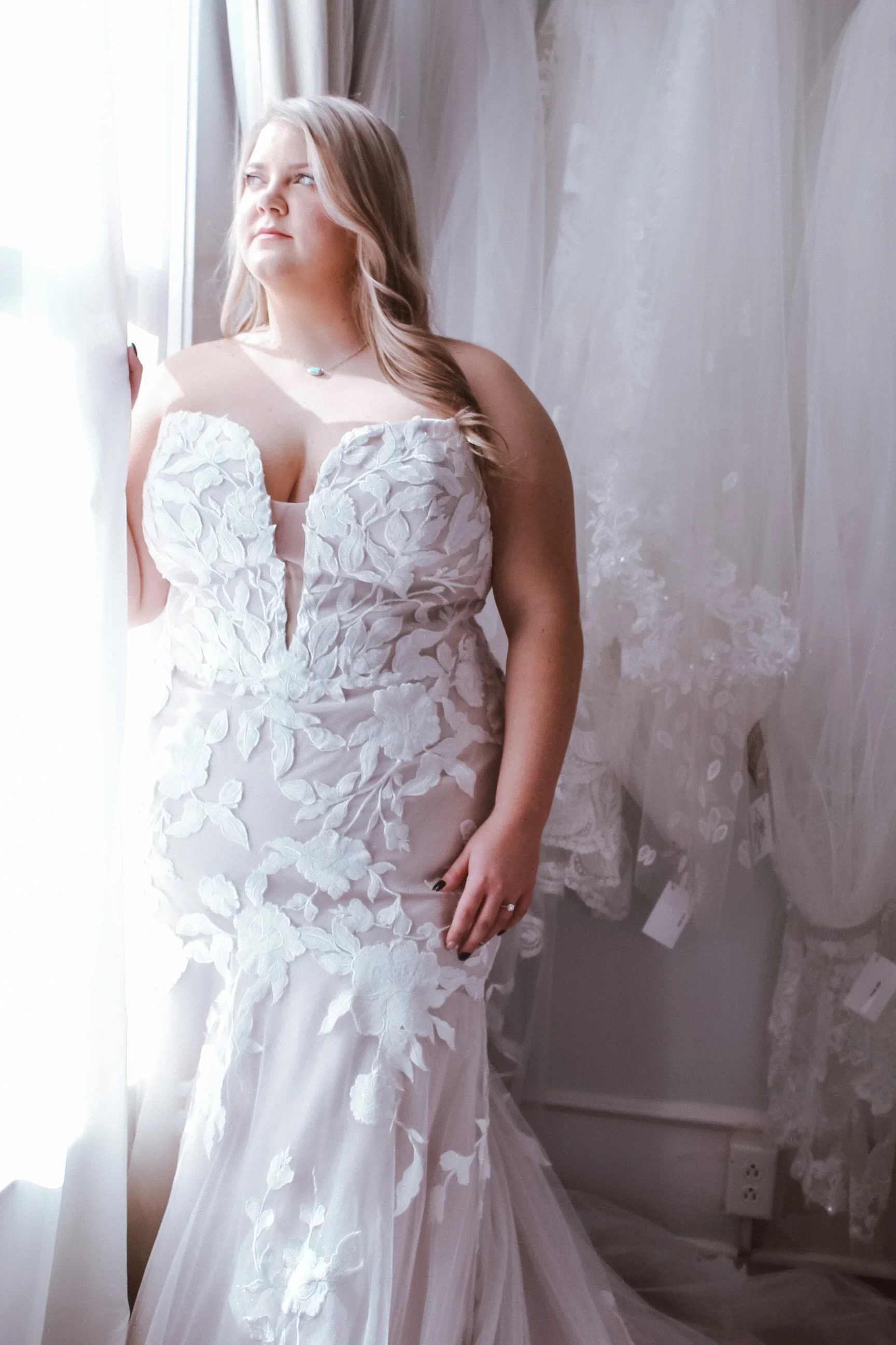 Bride wearing a strapless fit-and-flare wedding dress featuring floral lace appliqués and a deep plunging neckline, standing by a softly lit window surrounded by hanging gowns.