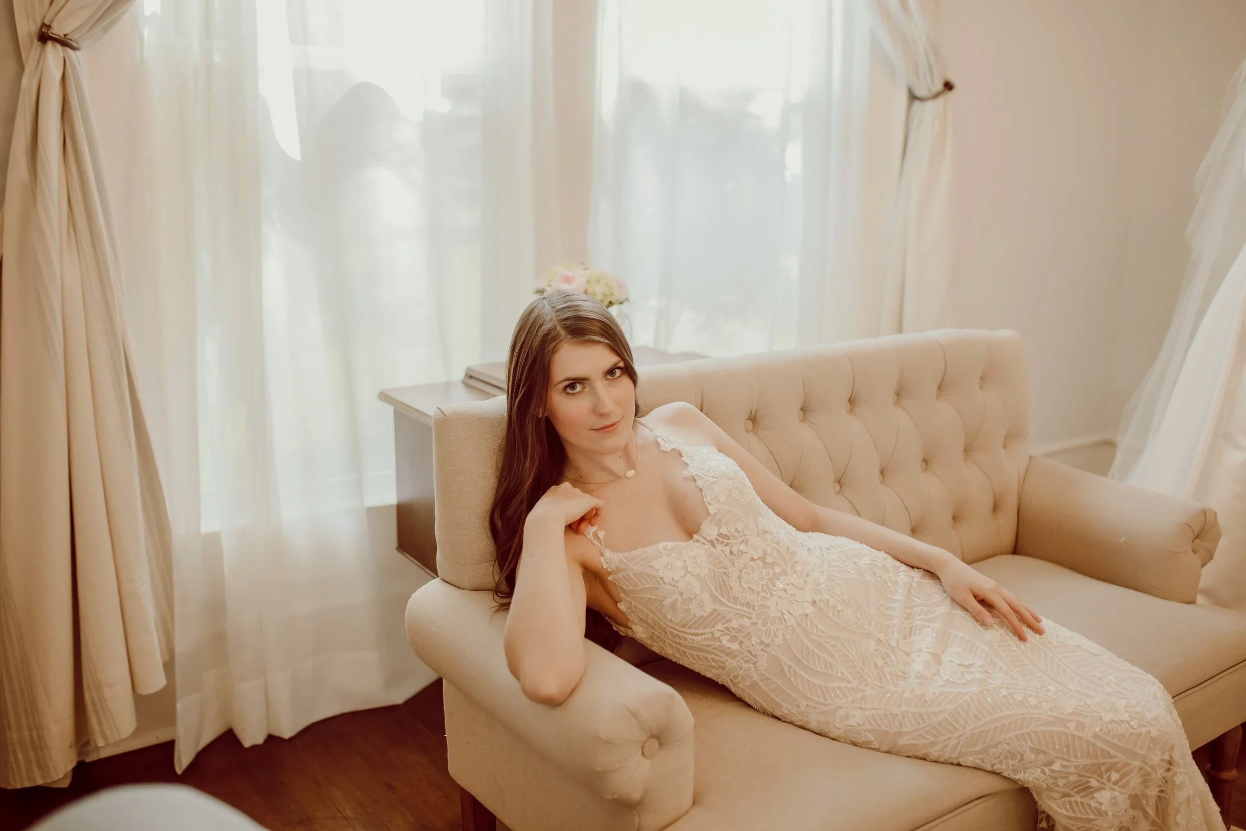 Bride reclining on a tufted beige sofa wearing a lace fit-and-flare wedding gown with delicate floral patterns, captured inside Atlanta Street Bridal Company’s softly lit bridal suite.