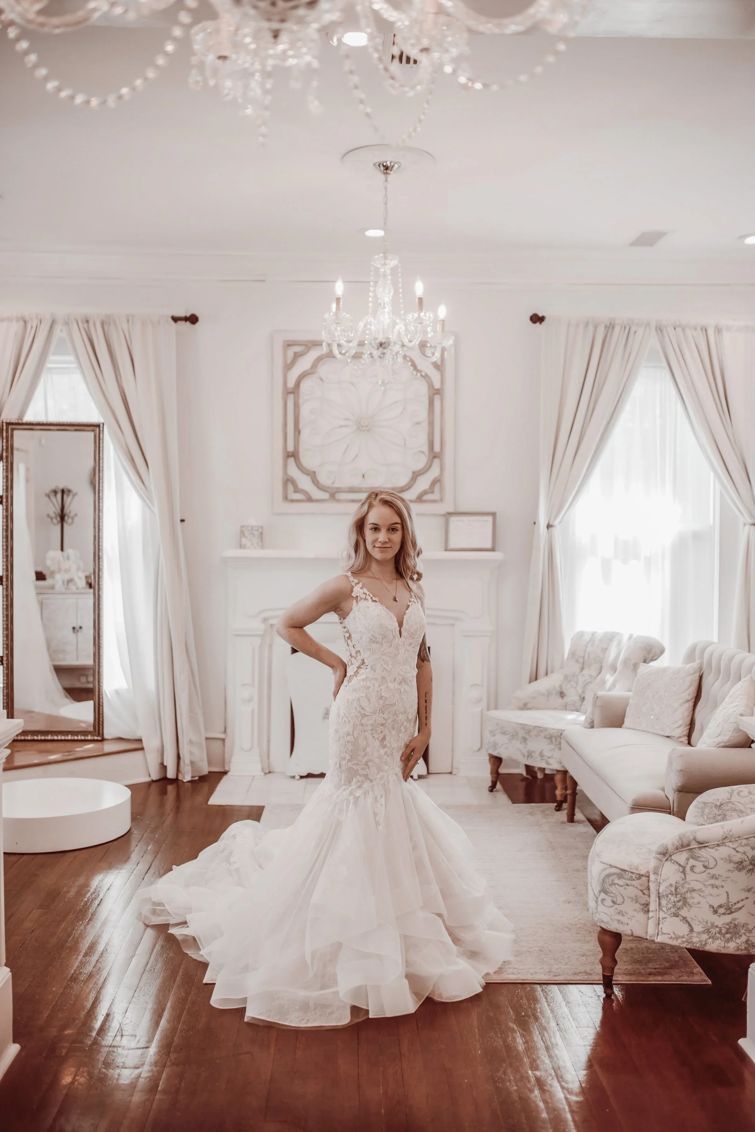 Bride standing elegantly in a bright bridal suite wearing a lace mermaid wedding dress with illusion straps, fitted bodice, and tiered tulle train under a chandelier.