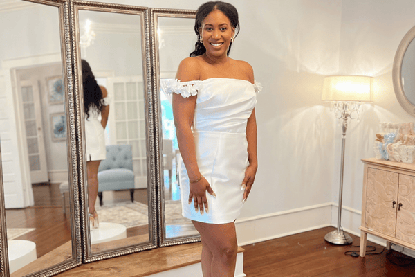 Smiling woman in a white off-shoulder dress stands in front of a large mirror, reflecting her image. The room is softly lit, creating a warm, elegant ambiance.