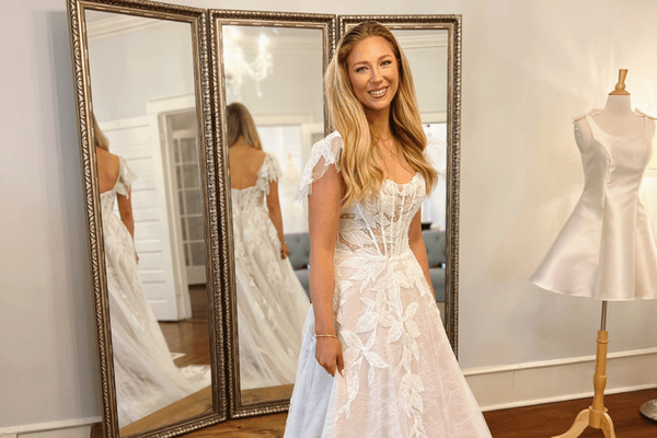 Bride wearing a lace A-line wedding dress with floral appliqué standing in front of mirrors at a bridal boutique.