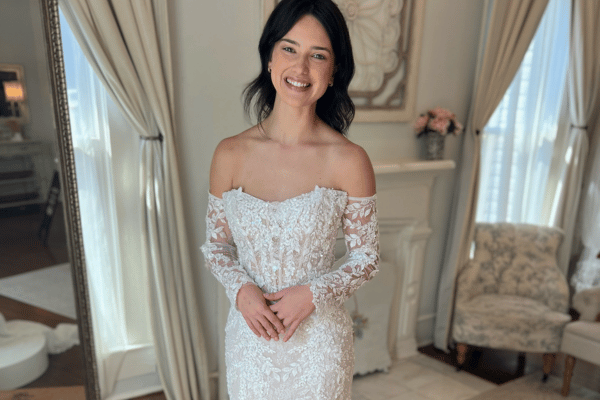 Smiling woman in a white lace off-shoulder wedding dress stands in an elegant room with beige curtains, an ornate mirror, and floral decor.