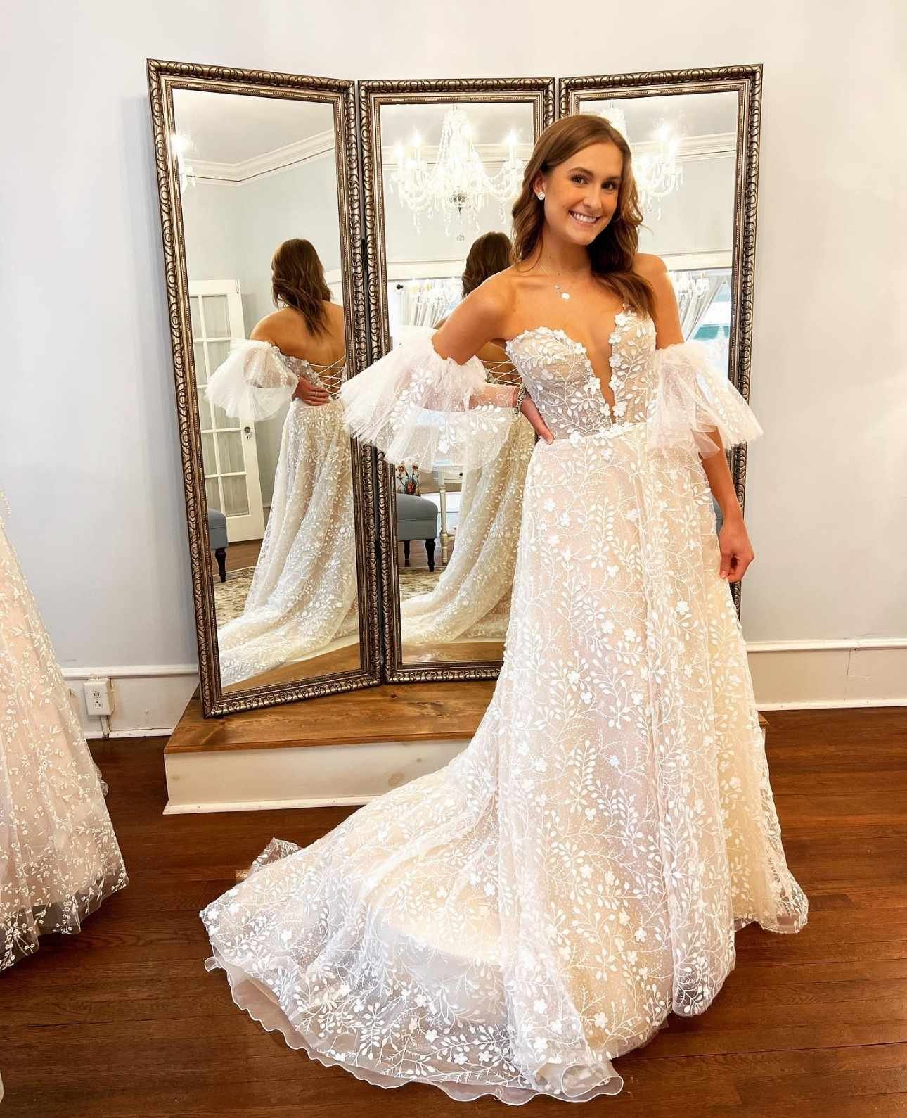 Bride wearing a floral lace wedding dress with a deep V neckline and sheer puff sleeves, standing in front of full-length mirrors inside the Atlanta Street Bridal showroom.