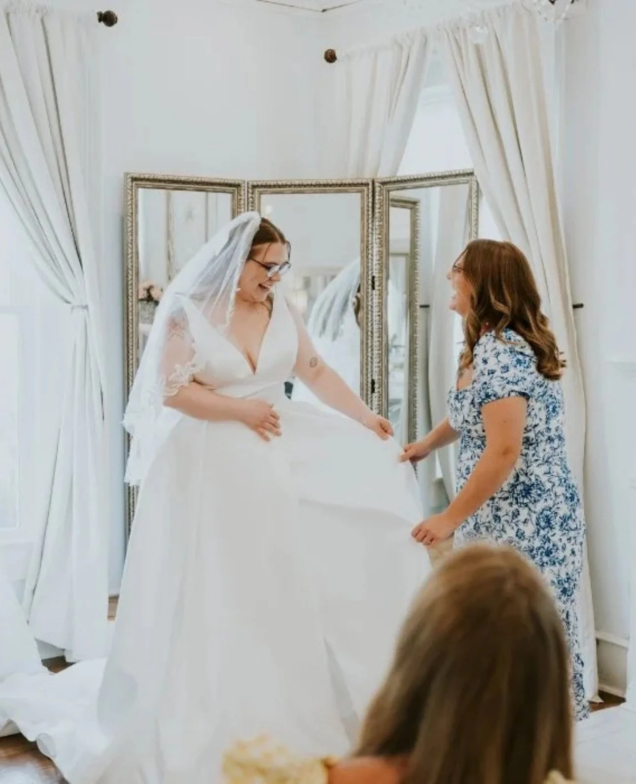 Plus size bride trying on a flowing wedding dress during a bridal appointment at Atlanta Street Bridal, assisted by a stylist in a bright fitting room with mirrors and white curtains.
