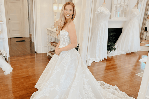 A smiling woman in a bridal shop tries on a strapless white wedding gown with a long, lace train, surrounded by mirrors and dresses.