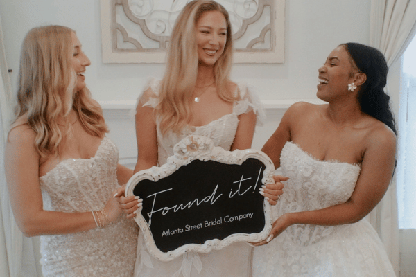 Three women in bridal gowns smile joyfully, holding a sign reading "Found it! Atlanta Street Bridal Company" in an elegant, warmly-lit setting.
