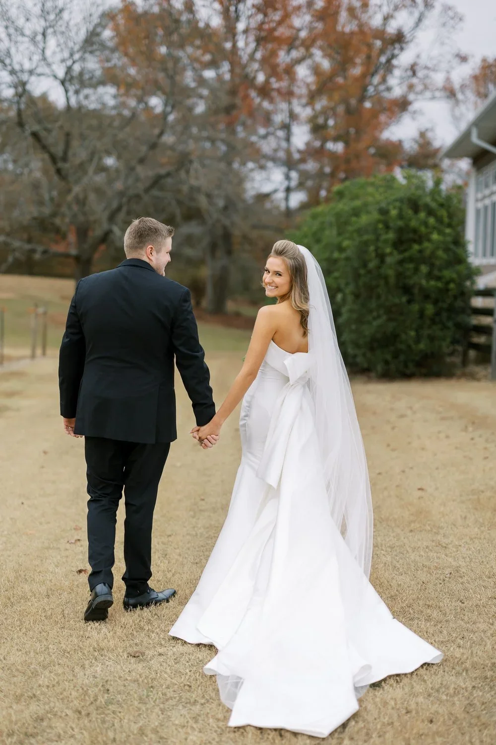 Bride in a strapless wedding dress featuring a dramatic bow back and long train, holding hands with her groom outdoors on a fall day, smiling over her shoulder.