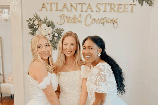 Three women in bridal gowns smile joyfully in front of a sign reading "Atlanta Street Bridal Company," with floral decor in the background.