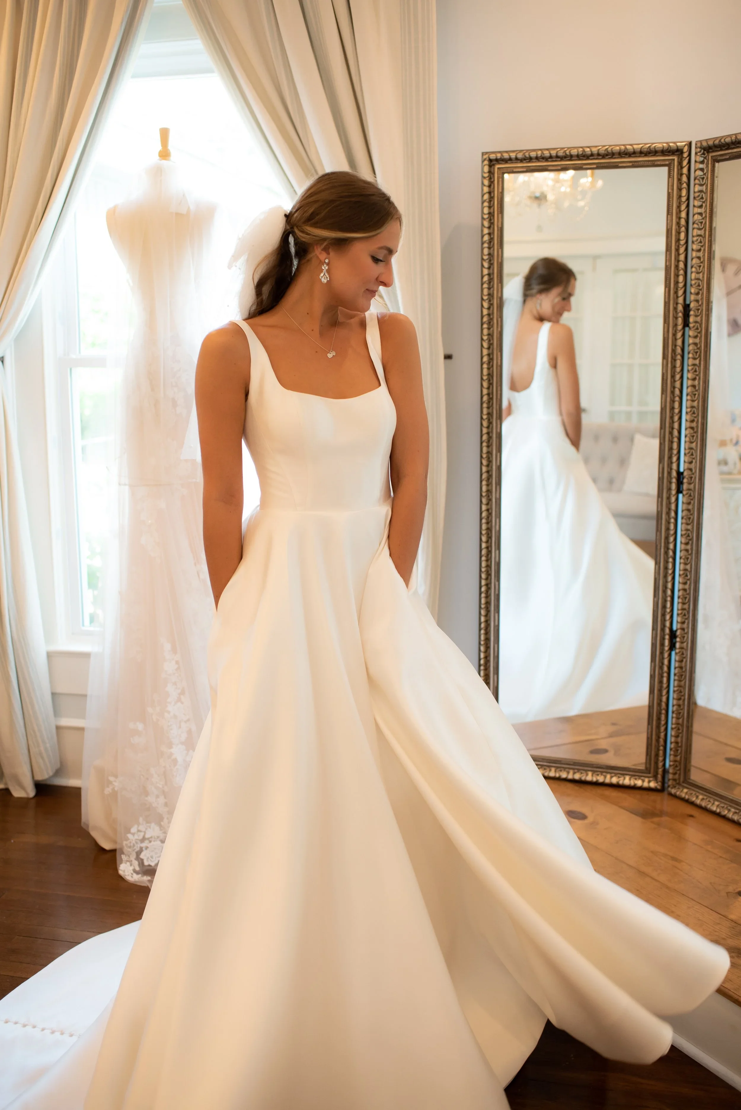 Bride wearing a timeless satin ball gown wedding dress featuring a square neckline, open back, and hidden pockets, posing gracefully in front of a mirror in an elegant bridal boutique.