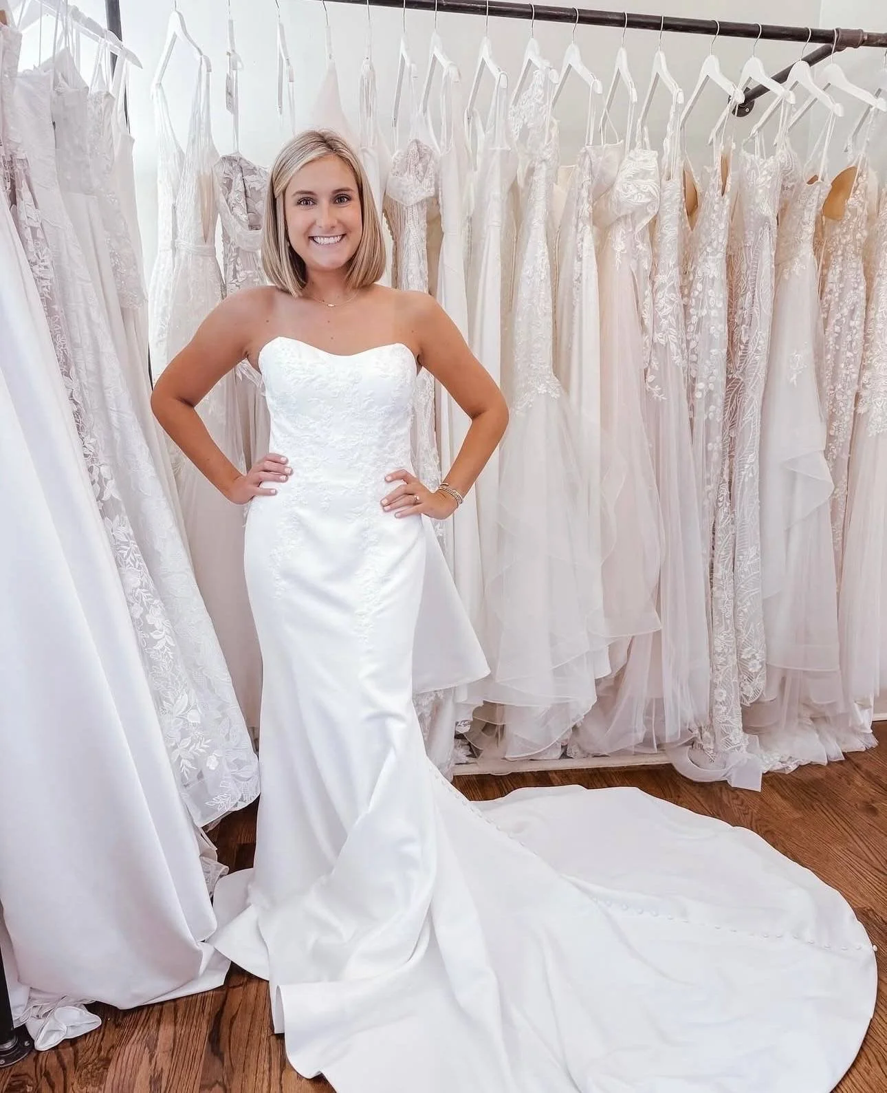 Bride wearing a strapless lace wedding dress with a fitted silhouette and chapel-length train, standing inside Atlanta Street Bridal surrounded by hanging bridal gowns.