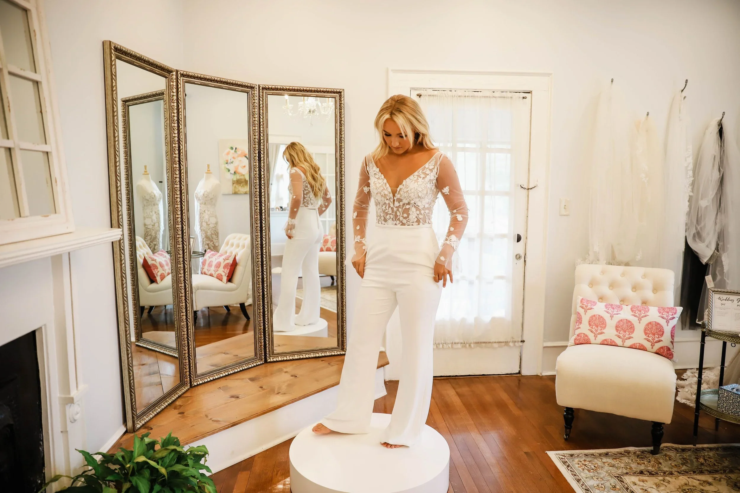 Bride wearing a modern two-piece bridal outfit featuring a sheer lace long-sleeve bodysuit with floral appliqués and high-waisted crepe pants, standing on a fitting pedestal at Atlanta Street Bridal Company