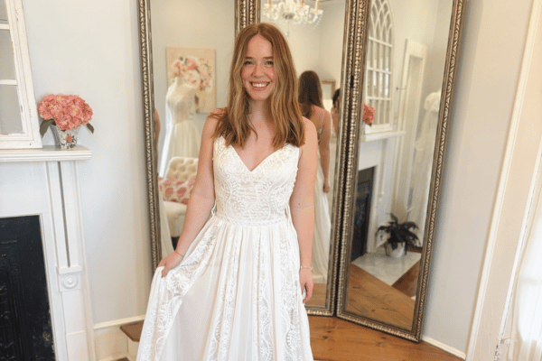 Smiling woman in a white lace wedding dress stands in a bright room with elegant mirrors and floral decor, conveying a joyful and celebratory mood.