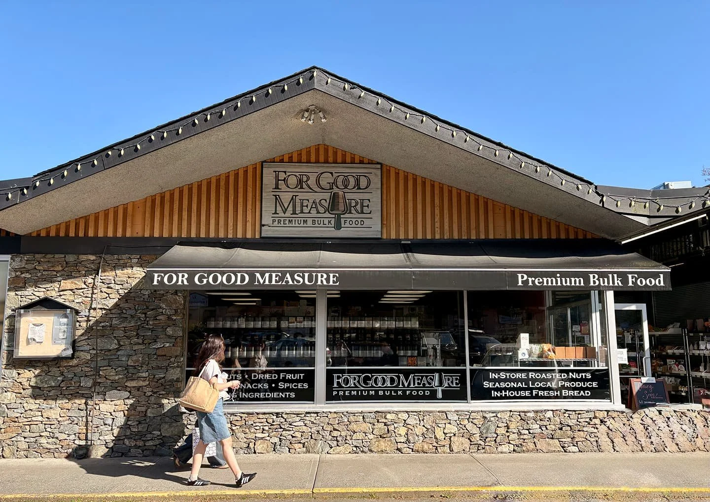 A woman walks past the stone exterior of For Good Measure, a premium bulk food store, on a sunny day.