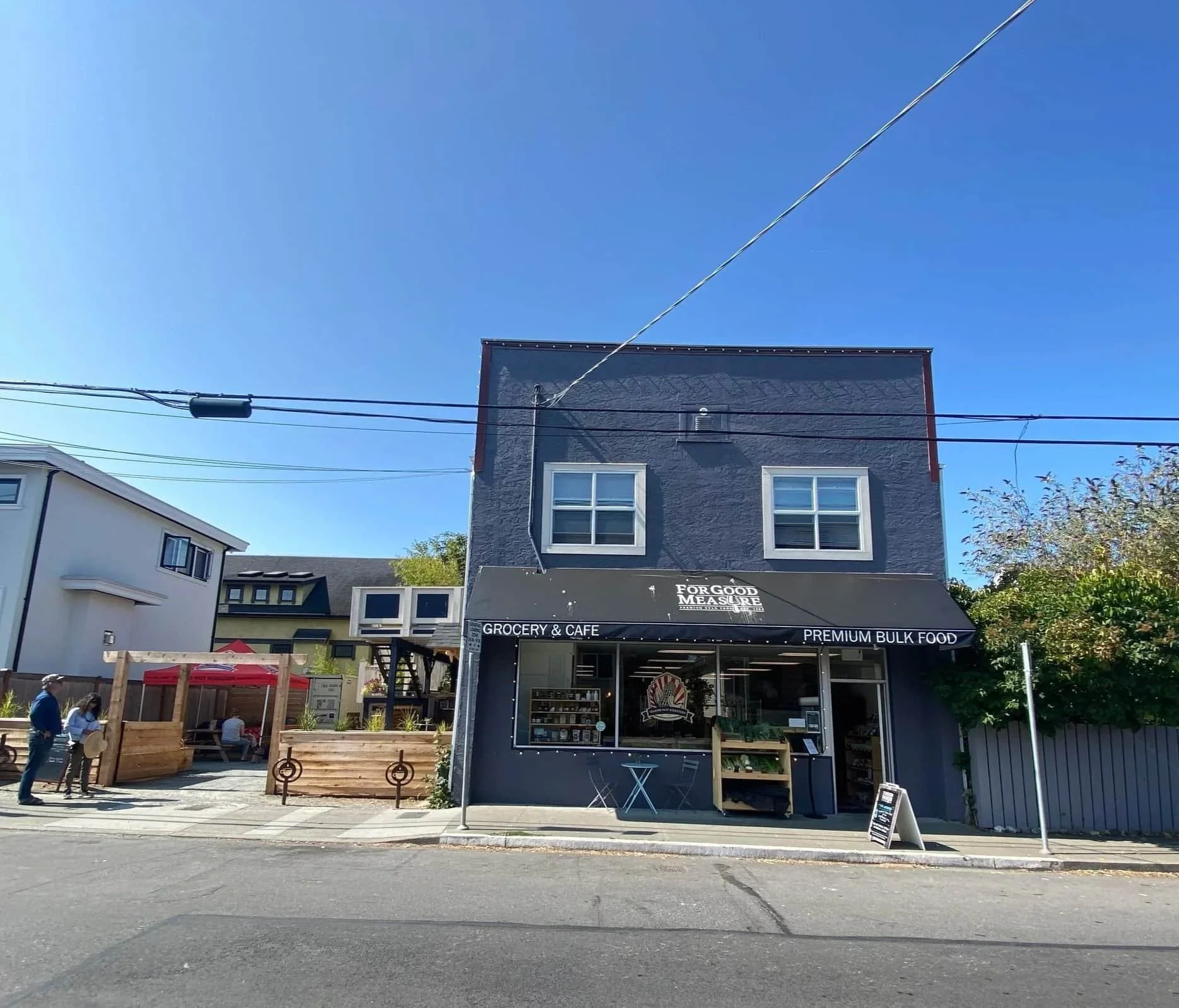 A two-story gray building with a grocery and café on the ground floor, outdoor seating, and clear blue sky overhead.