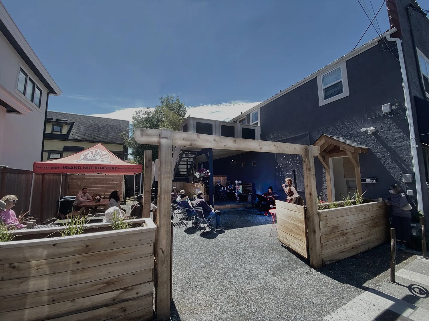 People enjoy a sunny patio with wooden planters, a canopy tent, and buildings—perfect for relaxing or catching James Bay live music.
