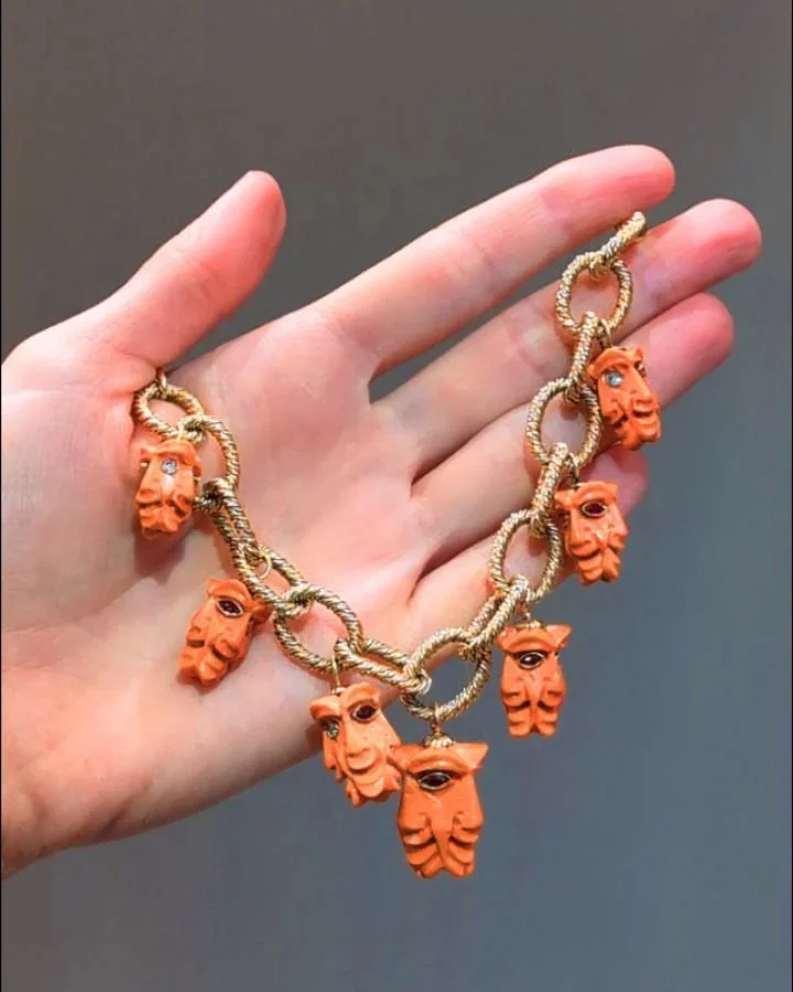 An 18kt gold charm bracelet of oval wire link design, suspending a series of seven gem set coral mask charms. Circa 1960. Available @simonteaklejewelry #charmbracelet #vintagecharmbracelet #coralcharms #vintagescharms #rubyanddiamond #simonteaklejewe