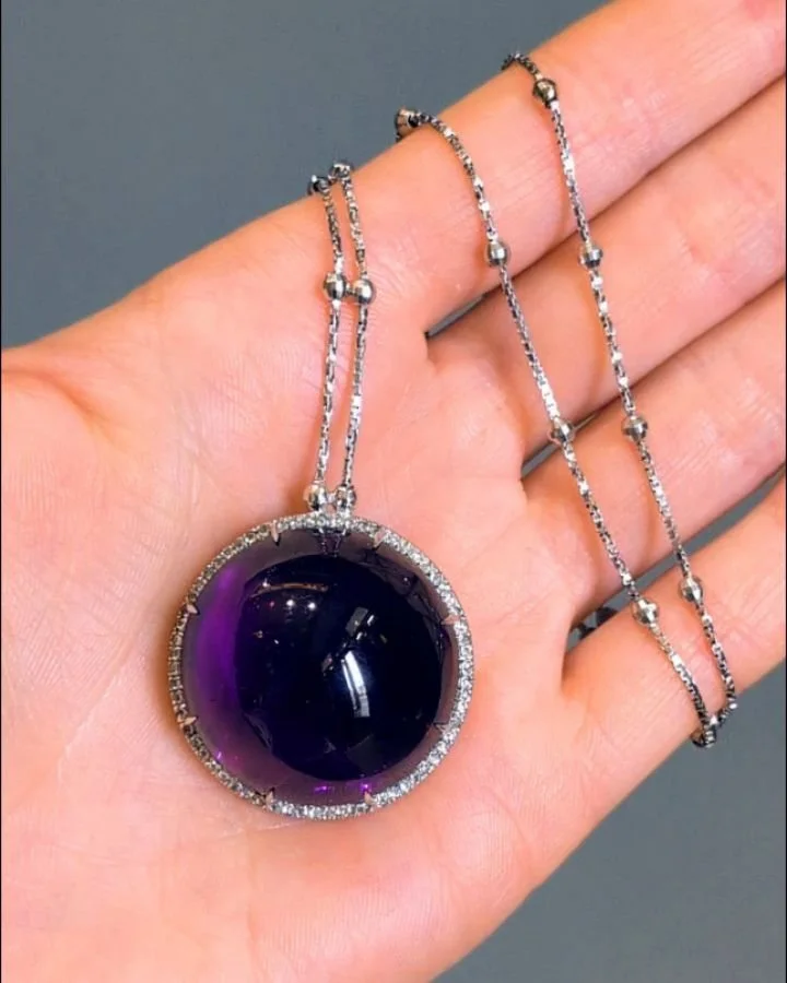 An amethyst and diamond pendant, the large circular cabochon amethyst of approximately 105.00cts with a micro pave diamond surround, mounted in 18kt white gold on a fine box link chain. Available @simonteaklejewelry #amethyst #amethystpendant #amethy
